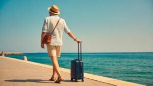 A man wearing a hat, light shirt, and shorts walks along a seaside promenade with a blue rolling suitcase, enjoying a sunny day by the ocean.