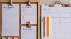 A corkboard with pinned sheets for weekly goals, a habit tracker, and focus notes sits beside a clipboard holding a blank weekly calendar. Three pastel pens (mint, yellow, peach) are clipped to the calendar.