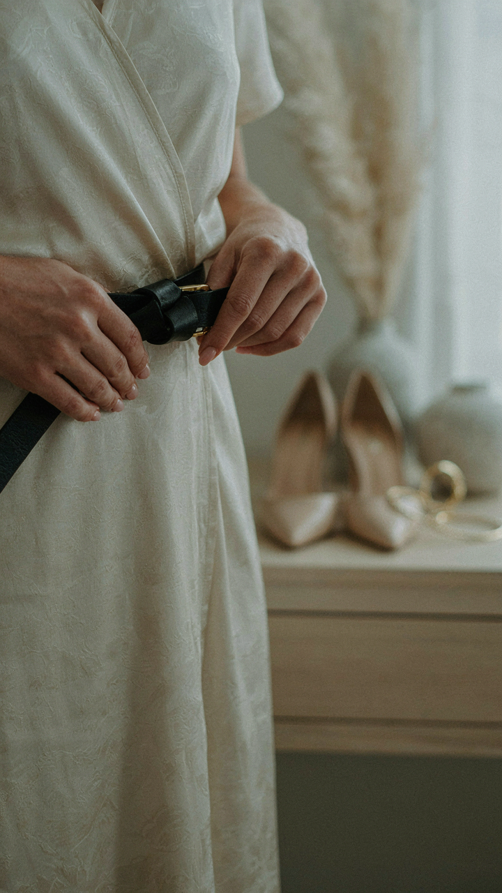 Hands tie a structured belt around a wrap dress, with shoes and jewelry nearby.