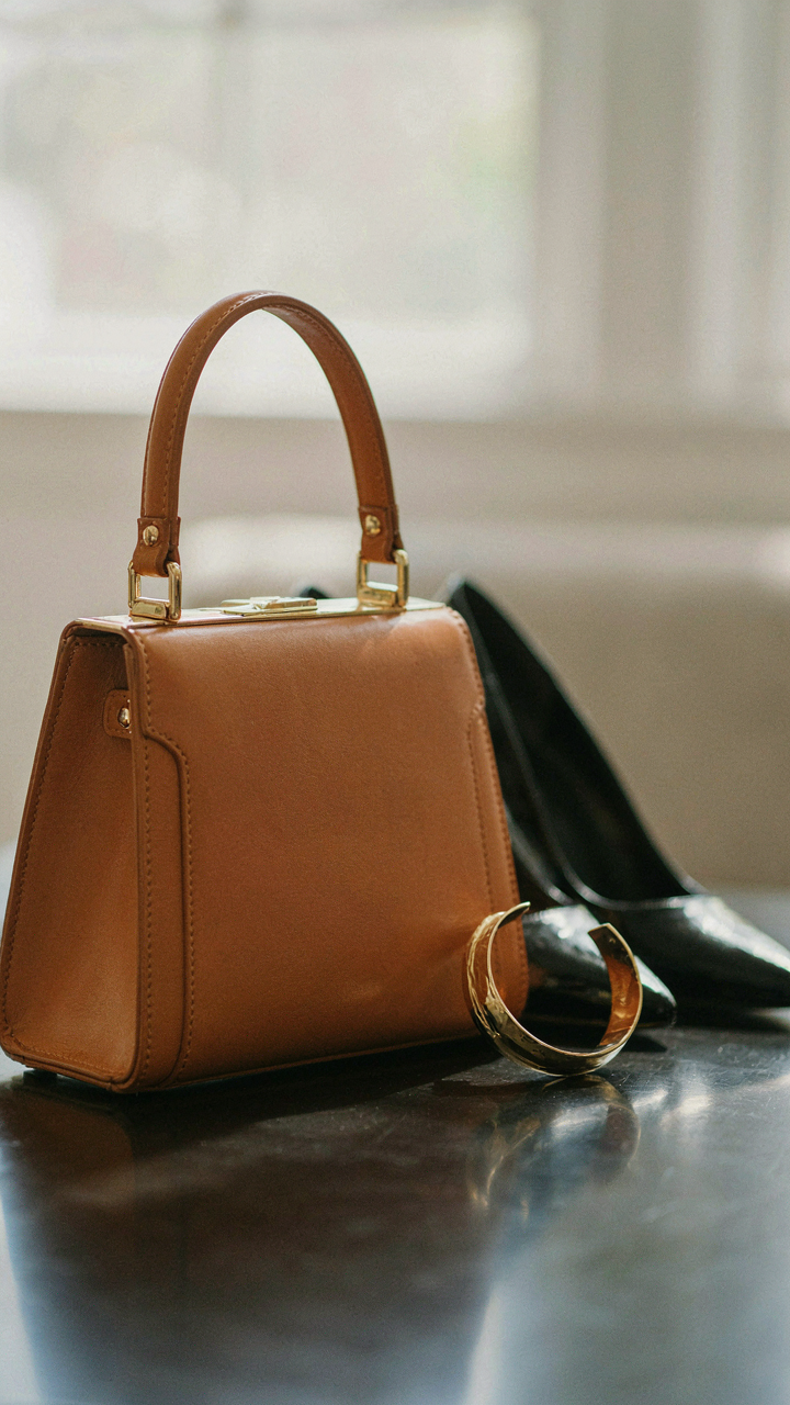 A structured leather handbag, gold bracelet, and black heels on a dark surface.