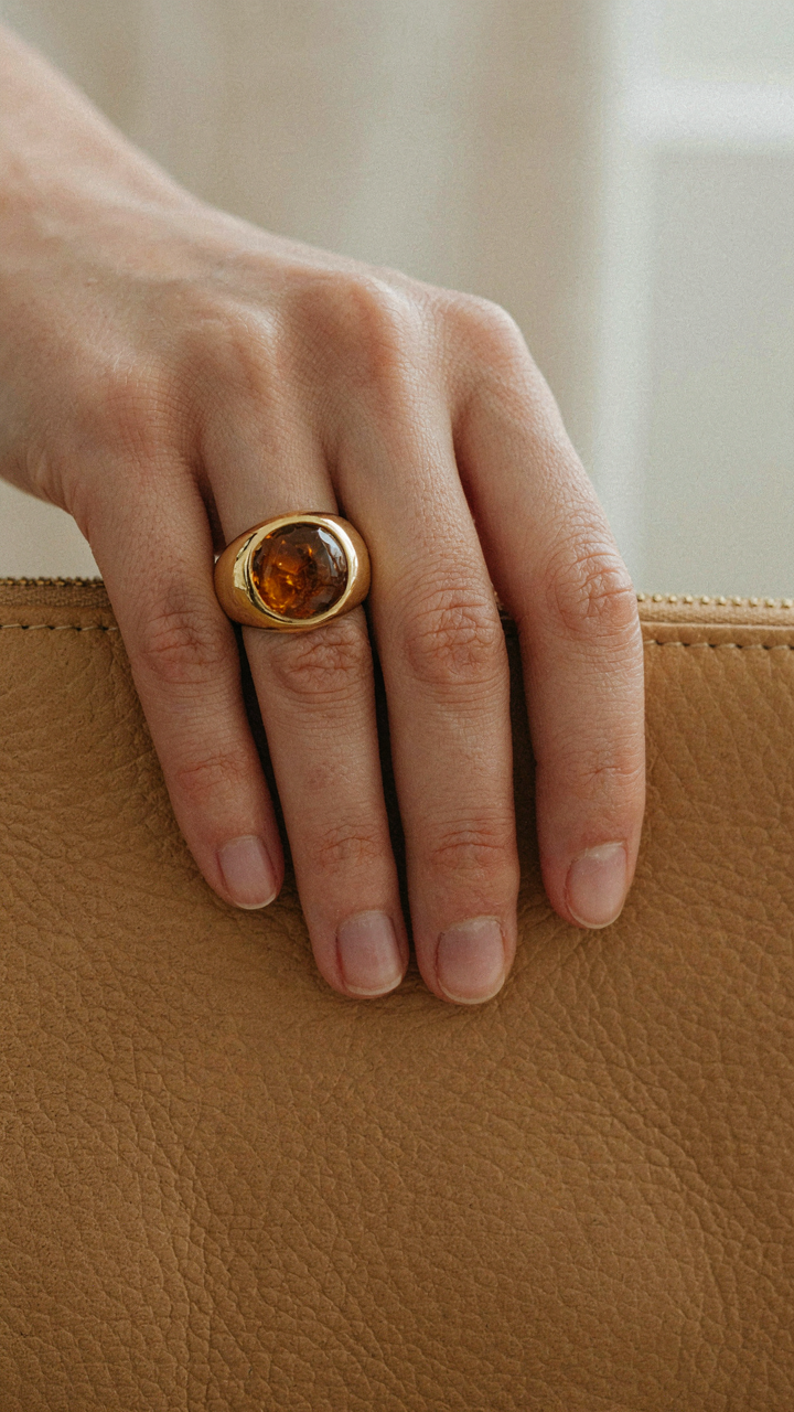 A hand wearing a single statement ring resting on a textured clutch.