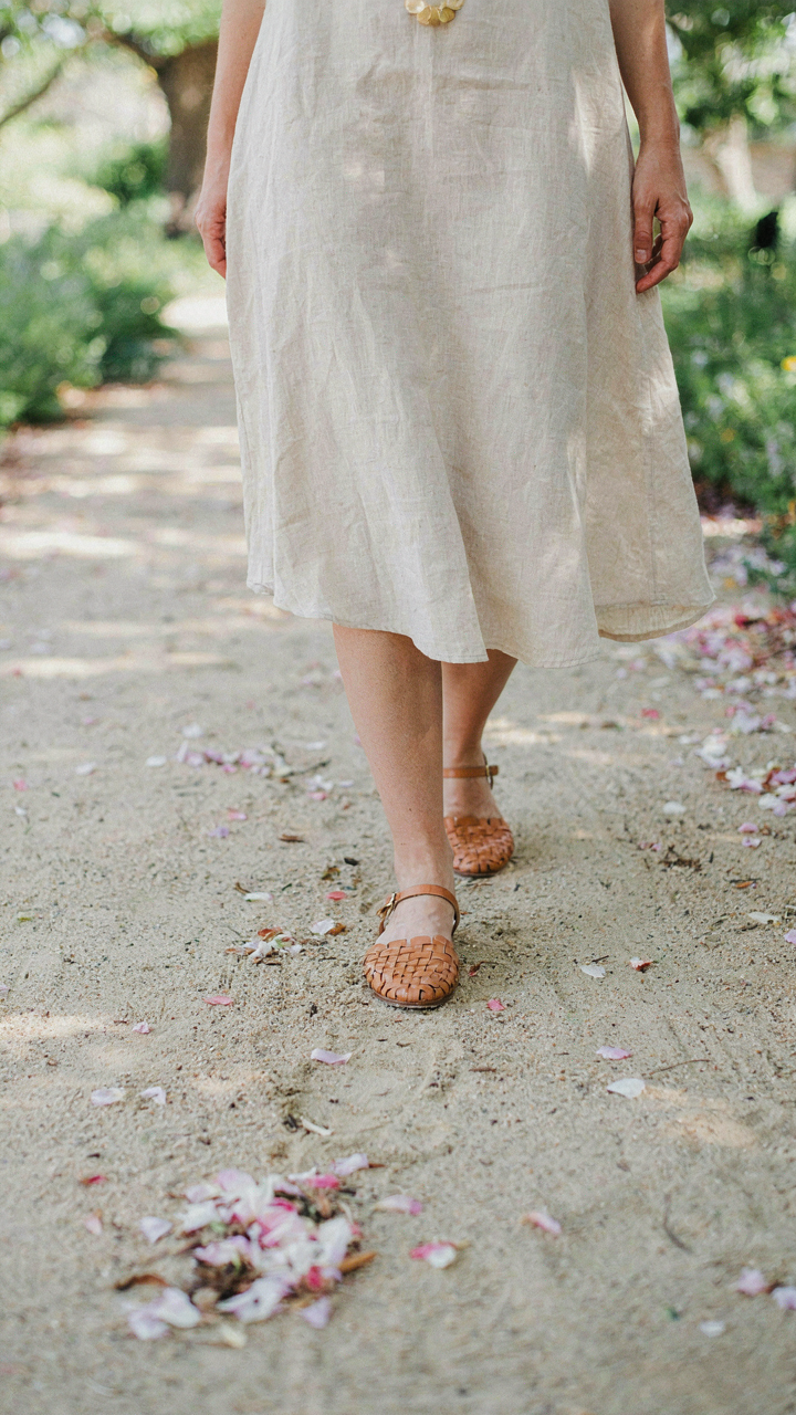 A person wears flat woven sandals on a sandy path with petals.