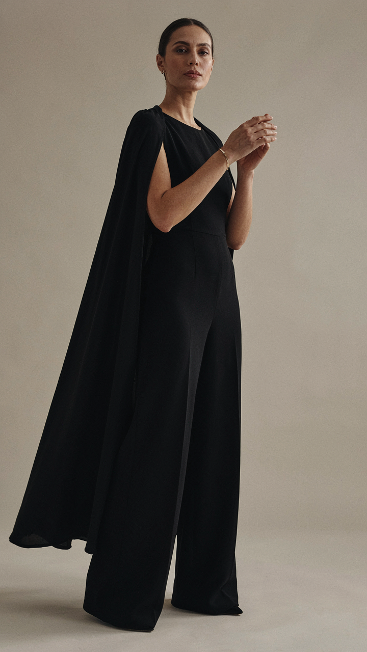 A single gold bracelet on a woman's wrist, complementing a dark formal jumpsuit with cape.