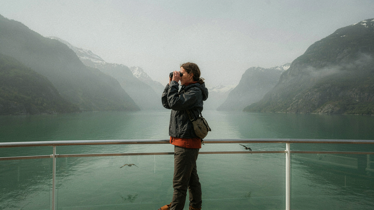 Woman in layered clothing looking at an Alaskan fjord from a cruise ship deck, binoculars in hand.