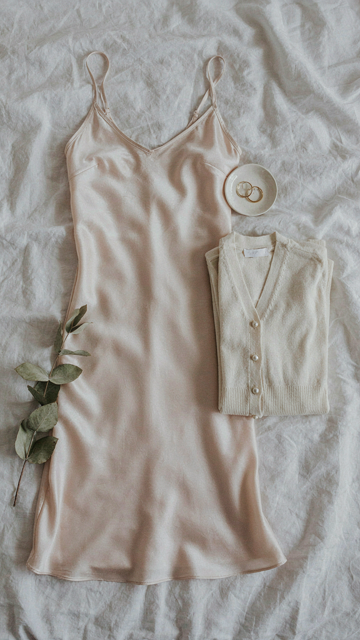 A silky slip dress and a folded silk blend cardigan lie on a bed.