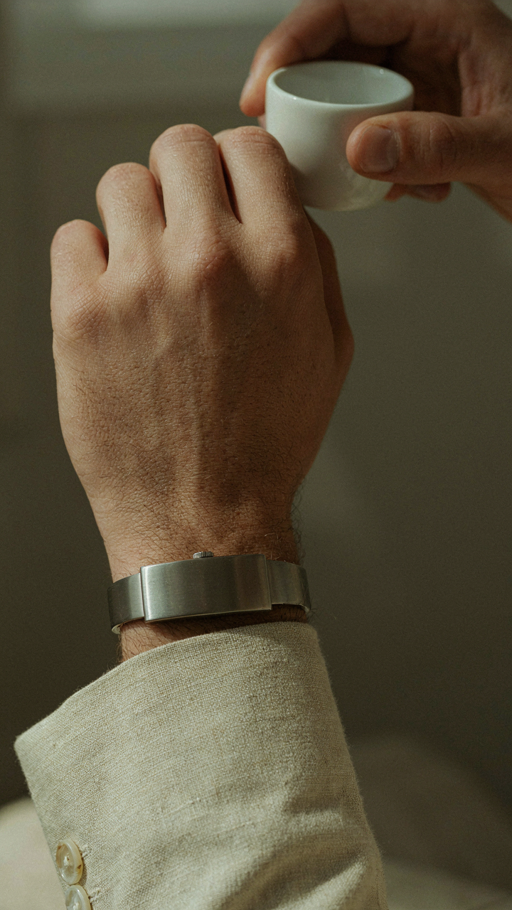 A man's wrist with a brushed metal watch and a linen blazer cuff.