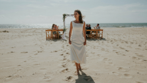 Woman in an elegant midi dress walking on the sand at a sunny beach wedding.
