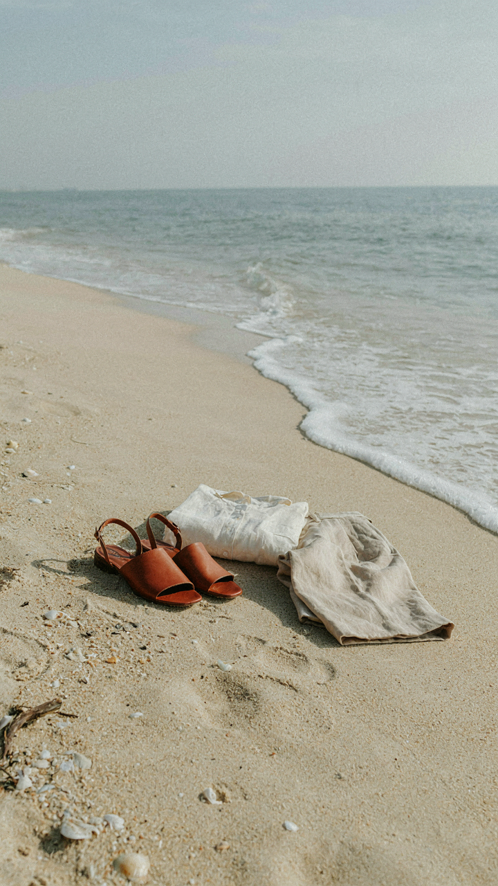 Leather slide sandals, a linen shirt, and trousers on a sandy beach.