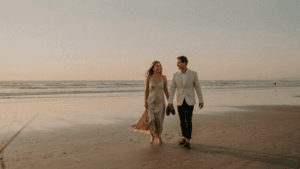 A well-dressed couple walks on a beach in elegant beach wedding guest attire at sunset.