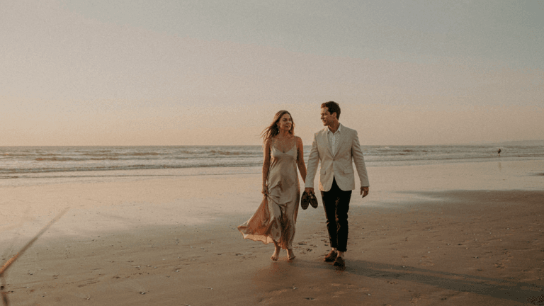 A well-dressed couple walks on a beach in elegant beach wedding guest attire at sunset.