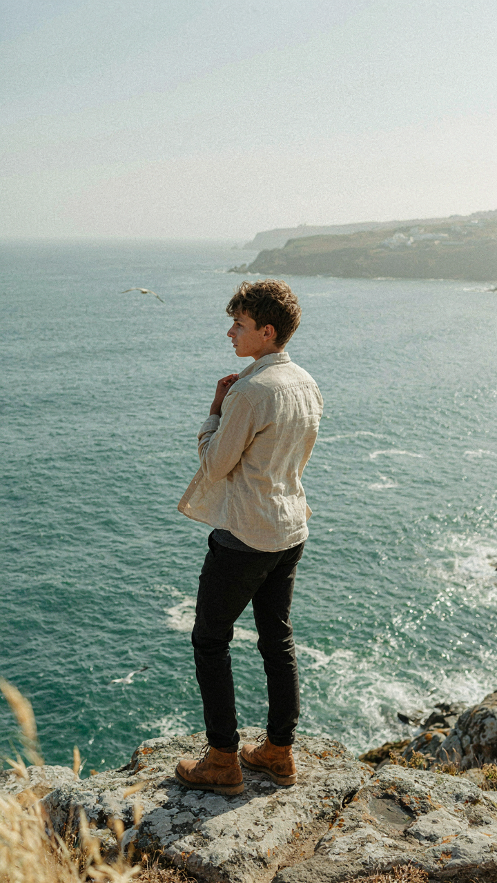 A person on a rocky coast adjusting a linen shirt worn over a merino base layer.