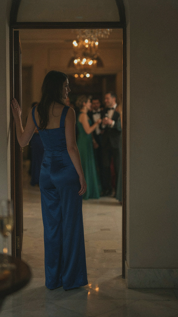 A guest in a sapphire blue satin jumpsuit observes a formal evening event.