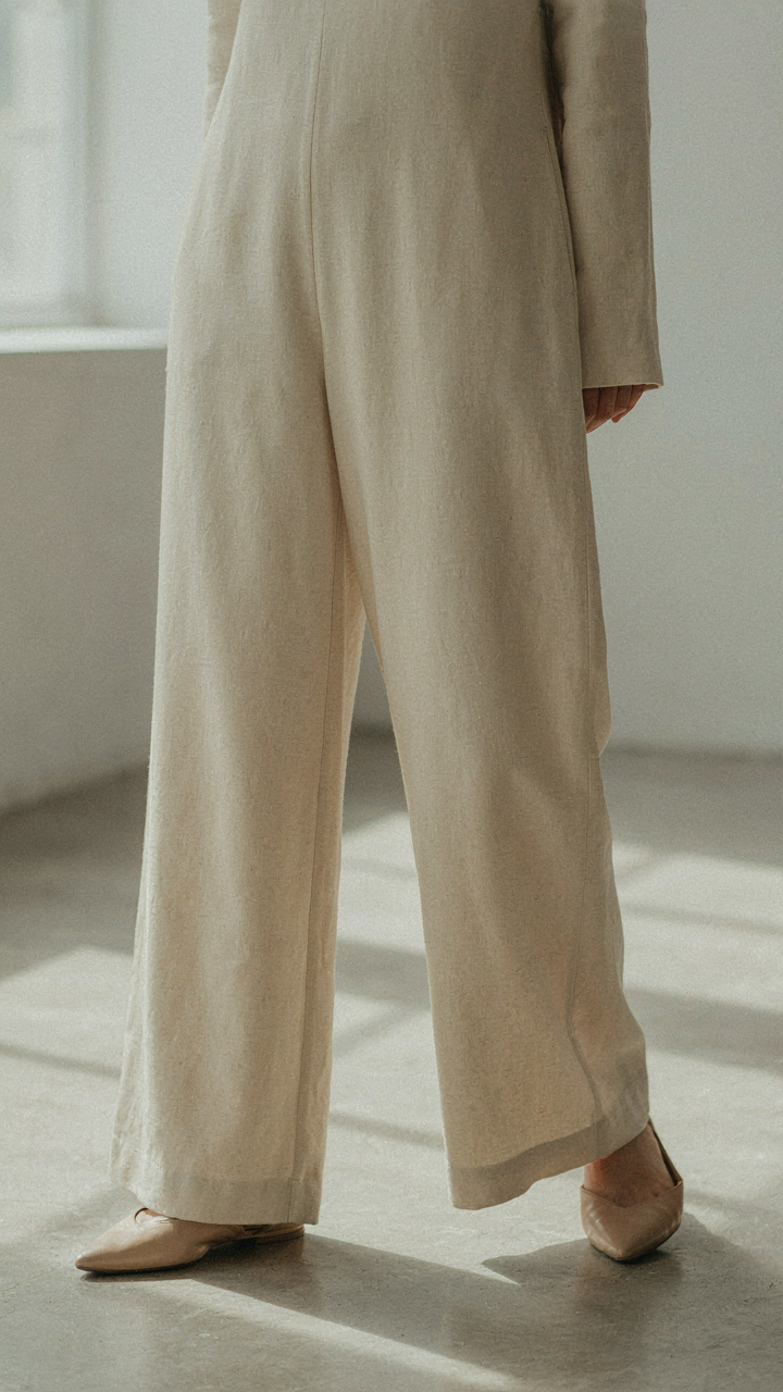 A woman wears a wide-leg linen jumpsuit with pointed flats on a concrete floor.