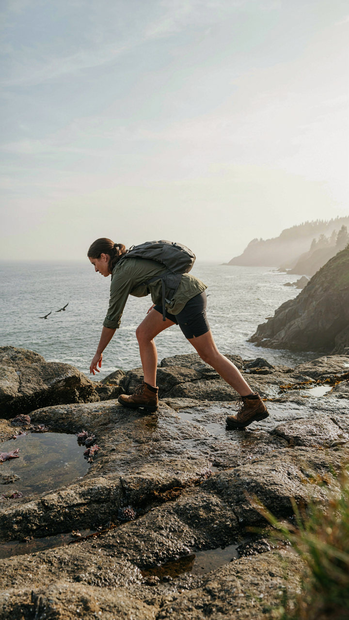 A person in practical hiking clothes climbs rocks to reach tide pools.