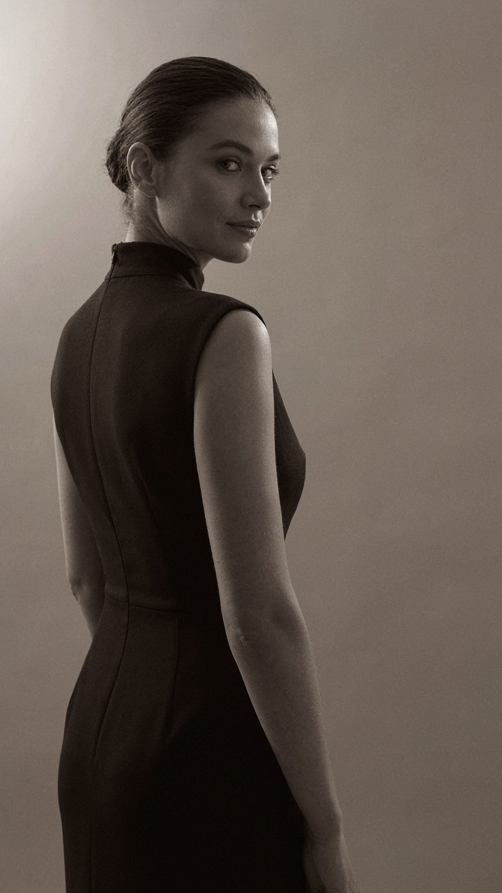 A woman in a dark high neck dress with perfect posture, seen from the back.