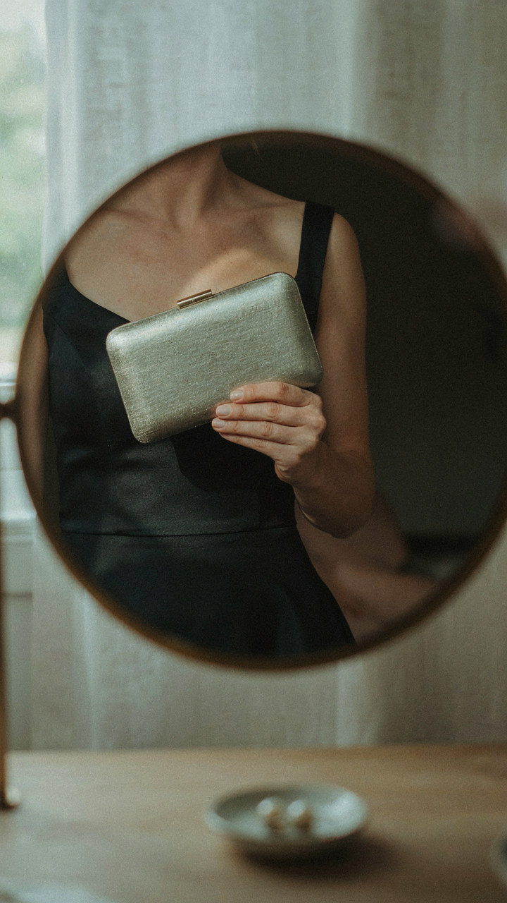 A hand holds a metallic clutch up to a mirror, reflecting a cocktail dress.