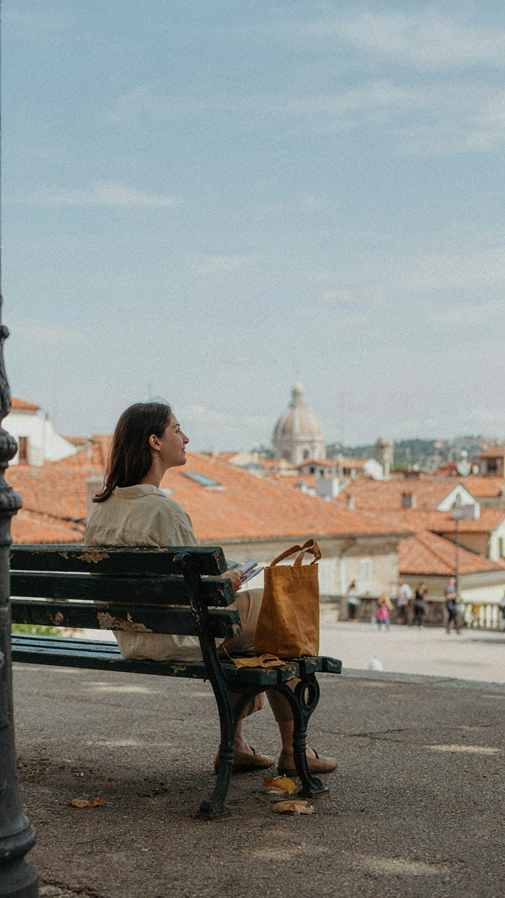 A calm traveler sits on a bench, looking at a city view with a small bag beside them.