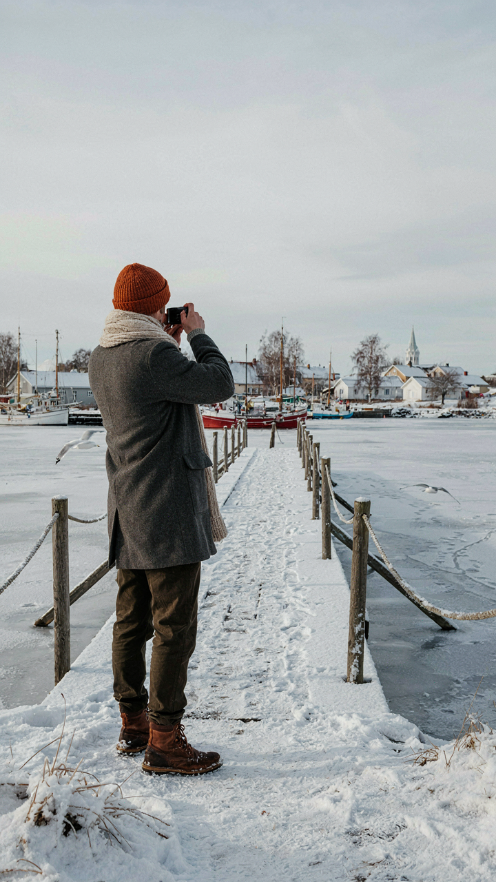 A person photographs a frozen harbor from a snowy bridge in a Scandinavian winter.
