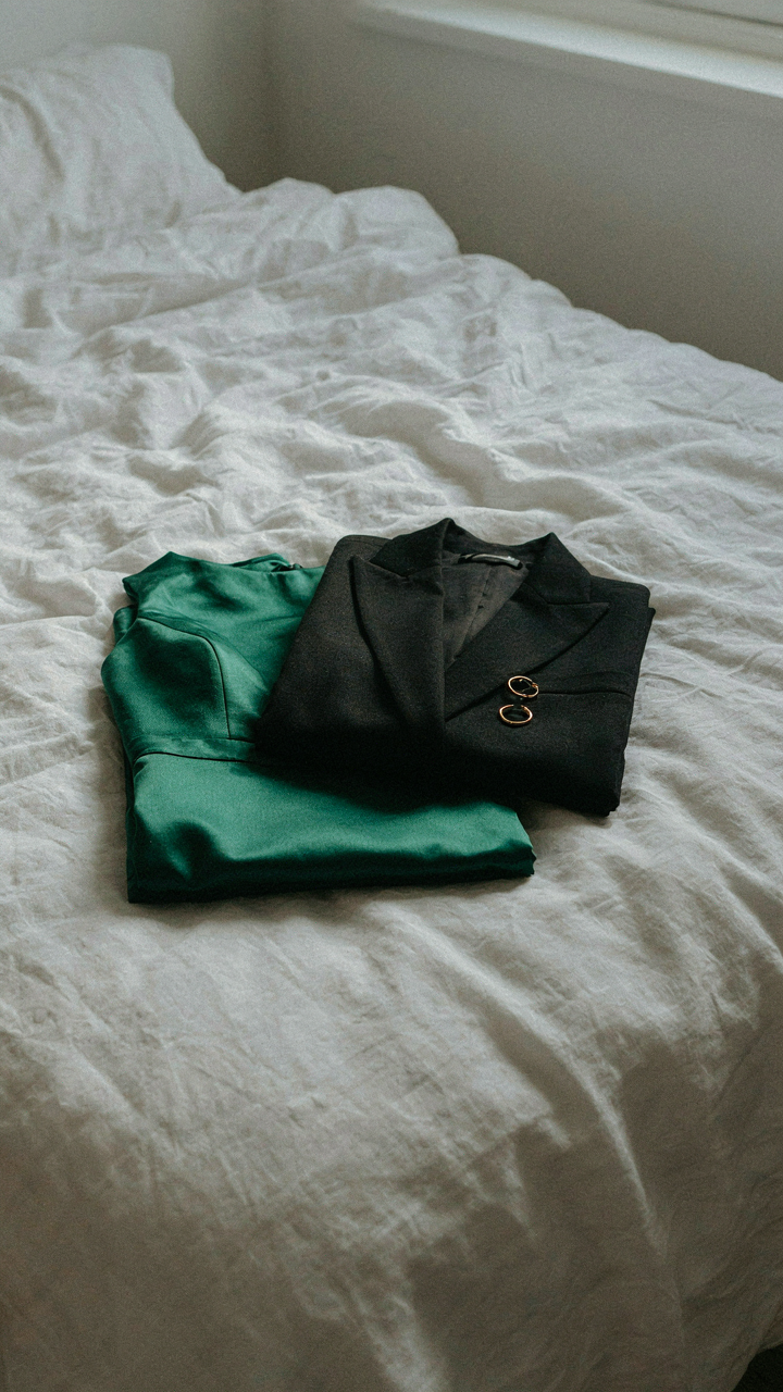 An emerald green formal gown with a black blazer lies folded on a bed.