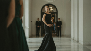 A woman wearing a black tie optional attire, a flowing black gown, stands at an event entrance.