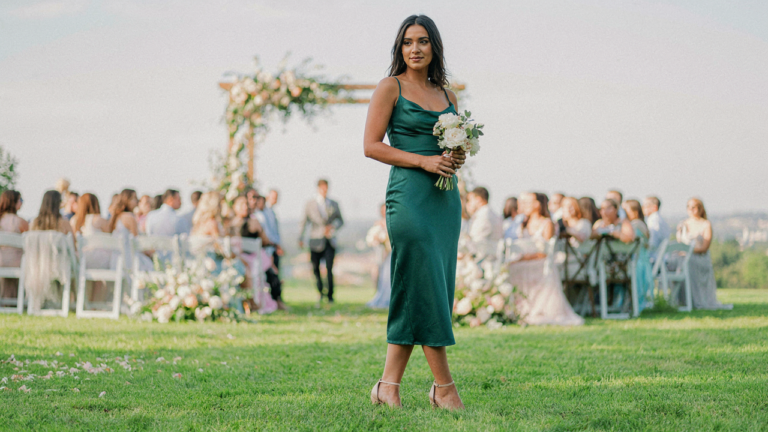 A woman in an emerald dress and block heels stands confidently at an outdoor wedding.