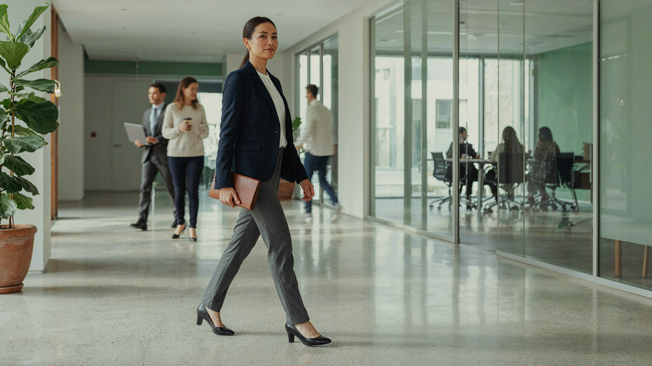 A professional woman in a sharp navy blazer walks through a modern, brightly lit office.