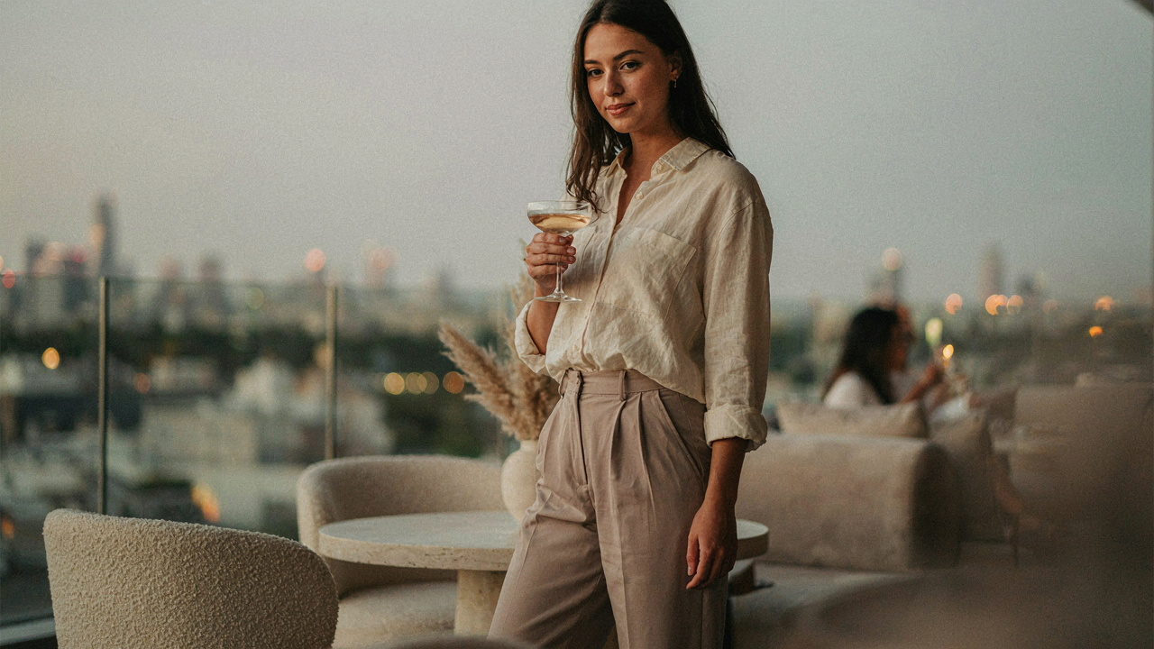 A person in stylish casual cocktail attire sips a drink at a relaxed evening party.