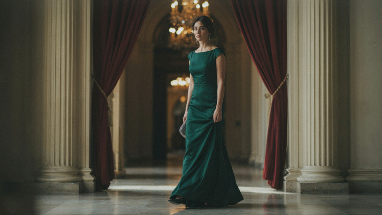 A woman in a beautiful jewel-toned gala dress walks into a grand ballroom.