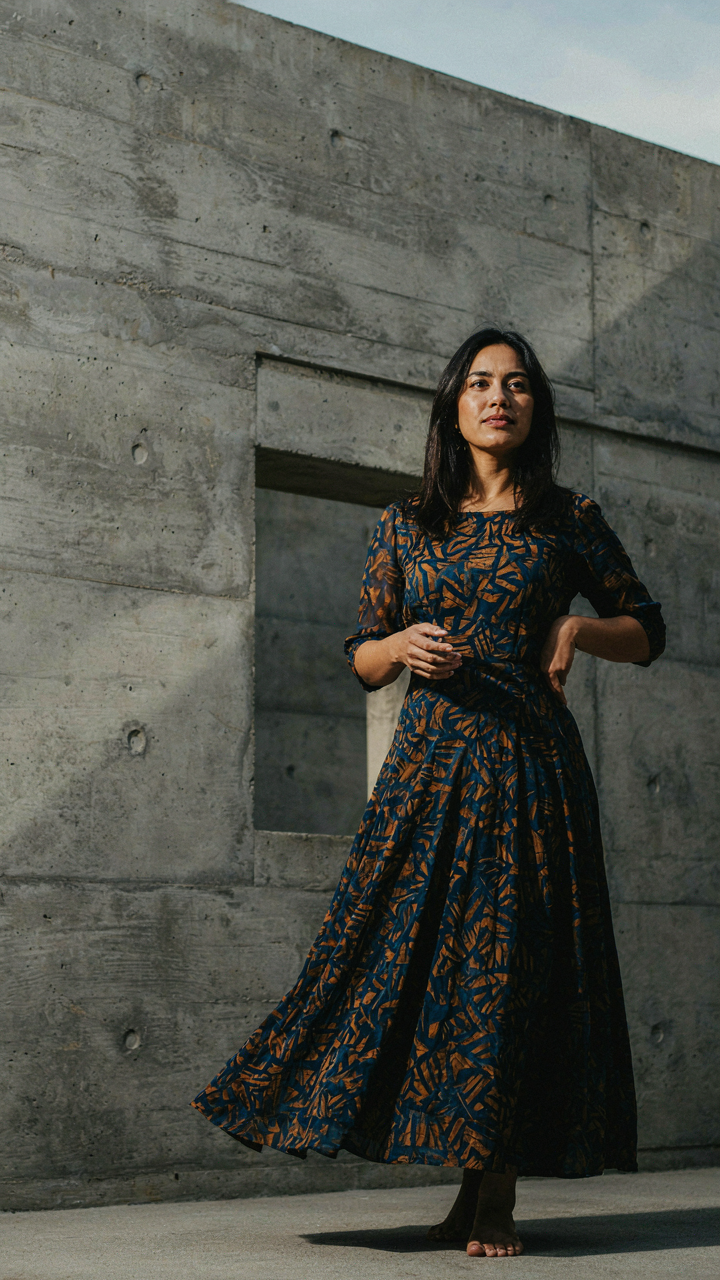 Woman in a flowing dress stands against a stark, geometric concrete building.