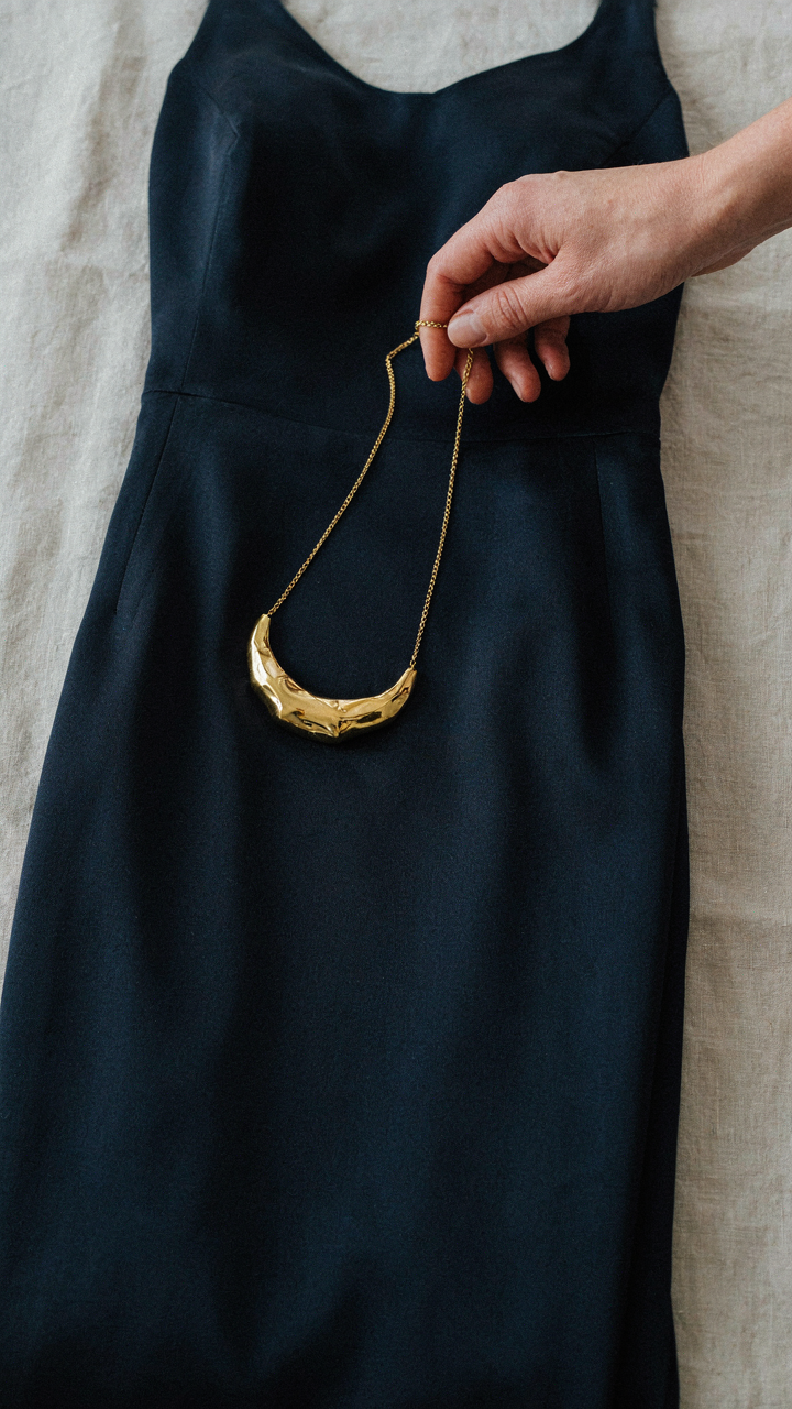 A hand places a statement necklace on a deep navy silk column dress.