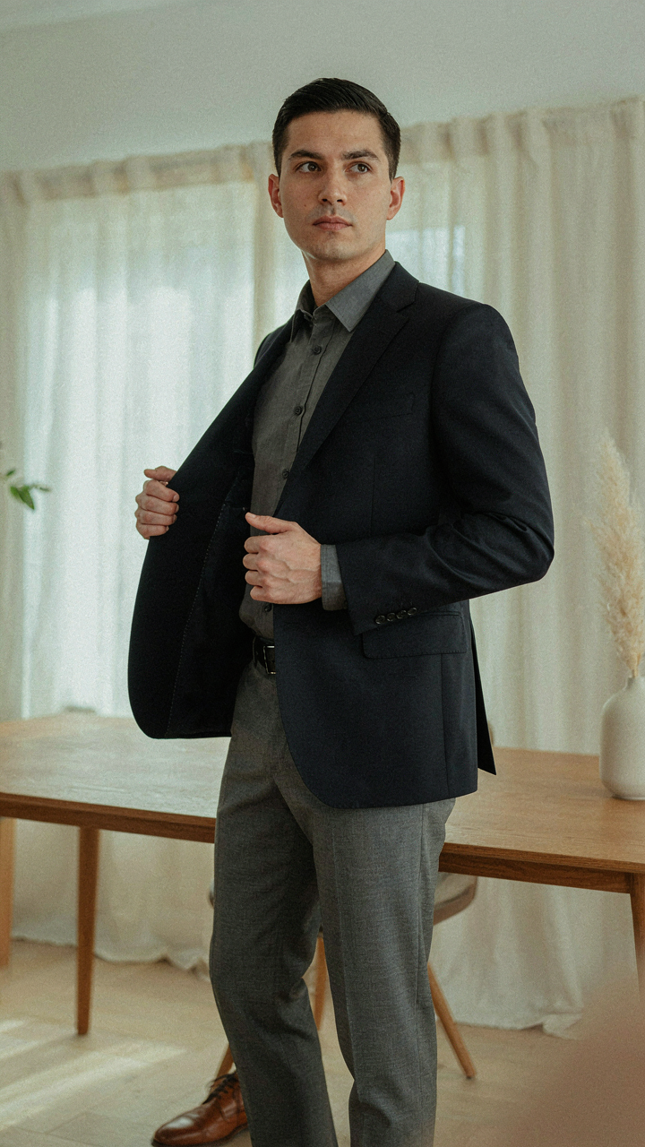 A person holds a dark blue blazer over tailored grey dress pants.