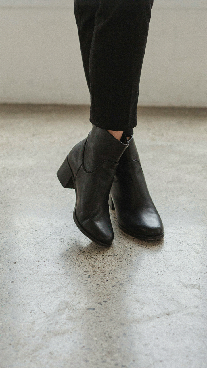 A person wears elegant, structured leather ankle boots on a concrete floor.
