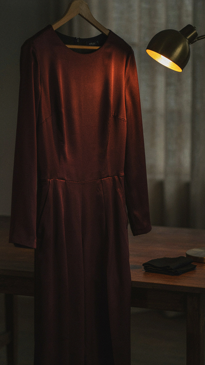 A burgundy satin jumpsuit on a hanger glows under a warm lamp.