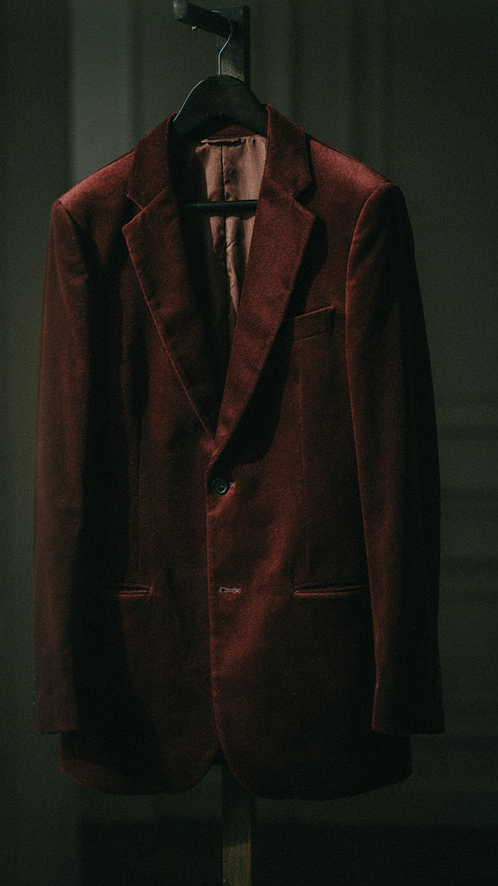 Deep burgundy velvet blazer hanging in a softly lit, dark room.