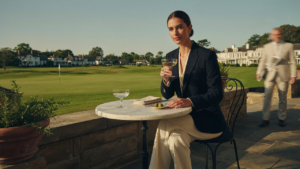 A woman in a navy blazer and cream slacks sips a drink at a sunny country club.