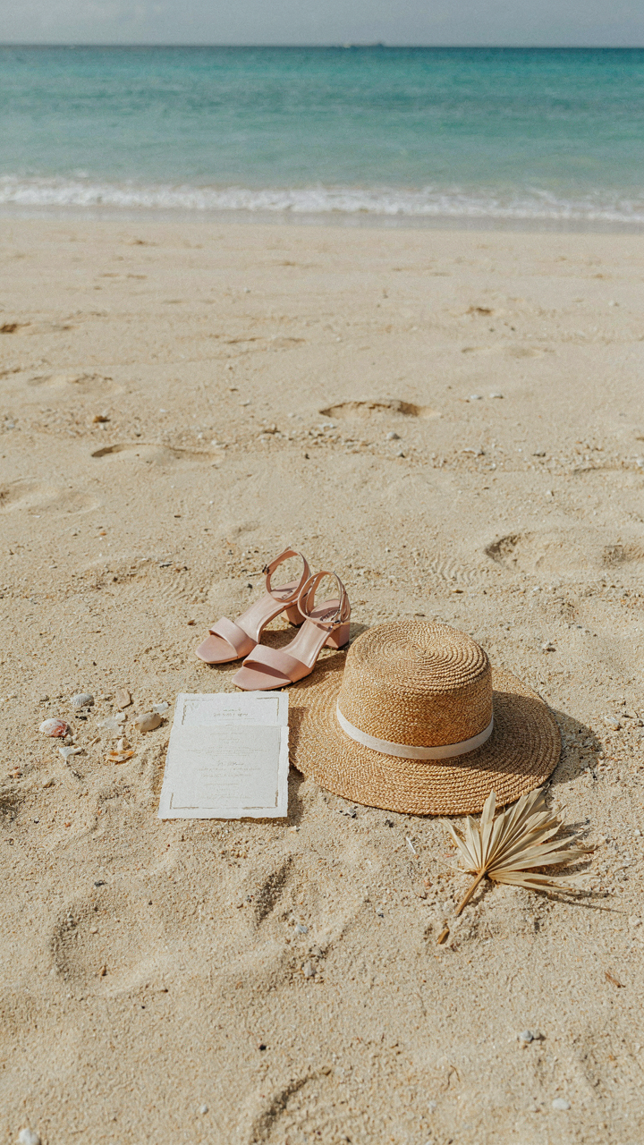 A cocktail attire wedding invitation sits on a sandy beach next to sandals and a hat.