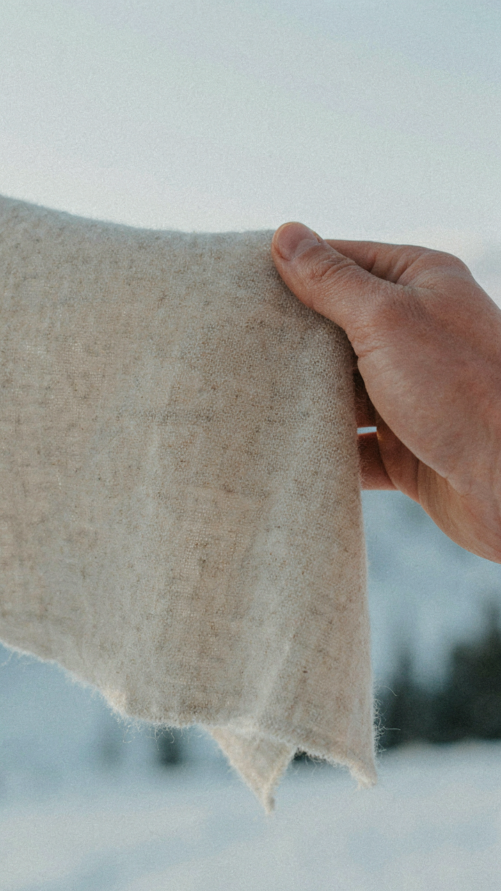 A hand holds up a piece of merino wool fabric against a snowy background.