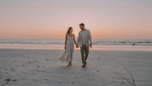 A stylish couple walks on a beach at sunset, dressed for a destination wedding.