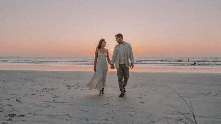 A stylish couple walks on a beach at sunset, dressed for a destination wedding.