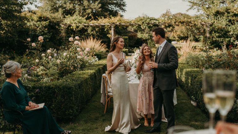 Wedding guests in elegant fascinators chat at an outdoor reception.