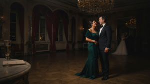 A formally dressed couple stands in a grand ballroom, ready for a winter wedding.