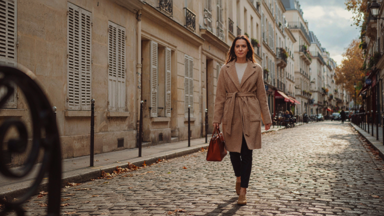 Woman in a camel coat and navy trousers walking on a Parisian street in autumn.