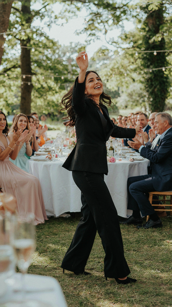 A woman in a black tuxedo jumpsuit dances freely at a wedding.