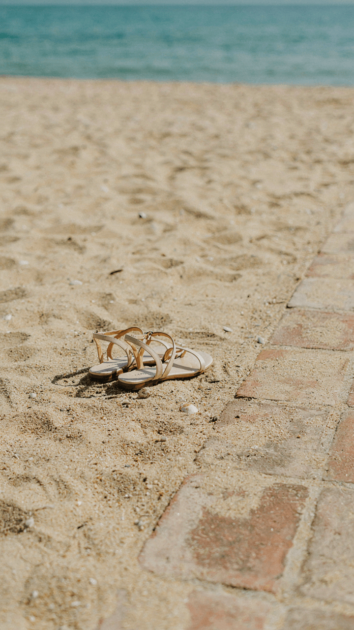 Elegant flat sandals sit next to sand and a tiled path.
