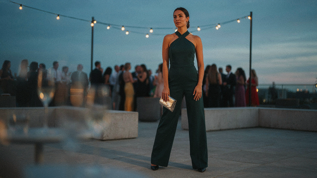 A woman wears a dark green halter jumpsuit, standing at an outdoor evening event.