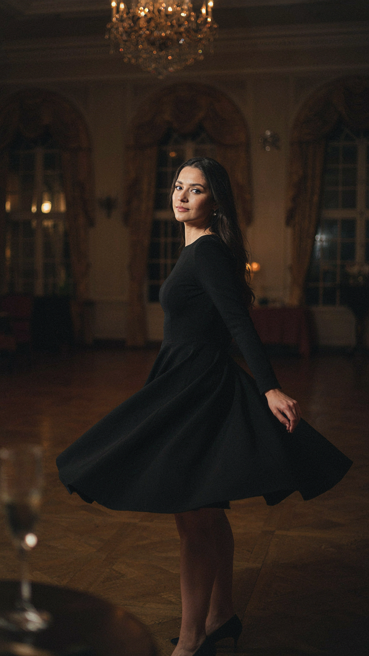 A woman in a black crepe A-line dress turns in a grand hotel ballroom.
