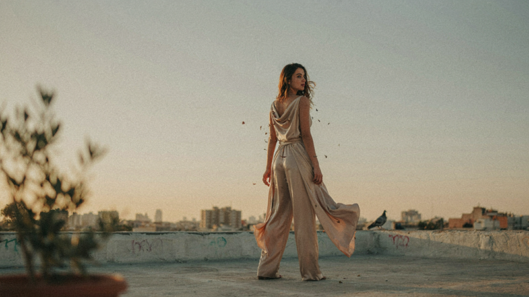 A person in a jumpsuit poses gracefully on a rooftop at sunset.