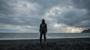 A woman in waterproof gear stands on a black sand beach during a windy day in Iceland.