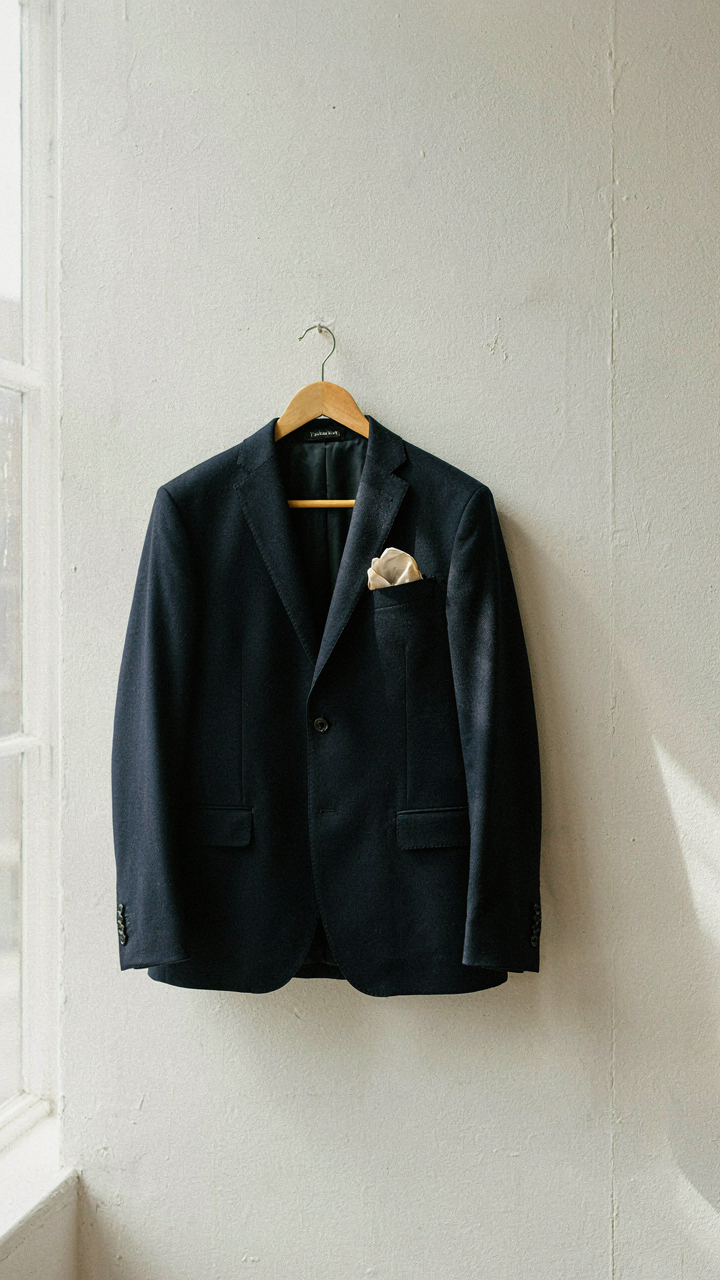 A tailored navy blazer hangs on a wall with a silk pocket square.