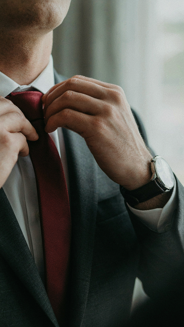 A man adjusts his silk tie on a dark suit, showing a simple watch.